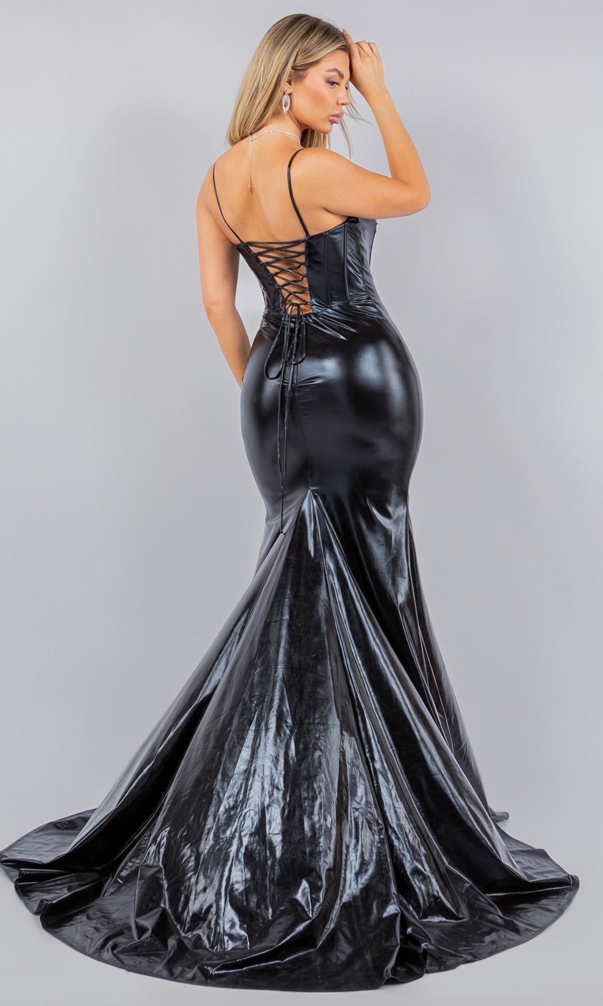 A woman models the Cinderella Couture 8092J Metallic Mermaid Prom Dress with a shiny black finish and lace-up back, standing against a light gray background and looking over her shoulder.